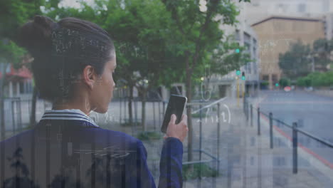 Animation-of-multiple-graphs-over-rear-view-of-biracial-woman-walking-and-using-smartphone