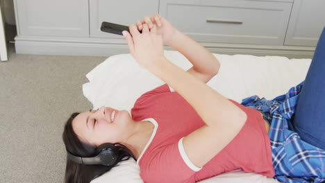 Glückliche-Biracial-Frau-In-Kopfhörern,-Die-Mit-Smartphone-Auf-Dem-Bett-Liegt,-Zeitlupe
