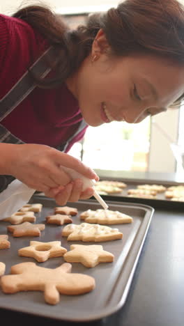 Vertical-video-of-happy-biracial-woman-decorating-chistmas-cookies-in-kitchen,-slow-motion