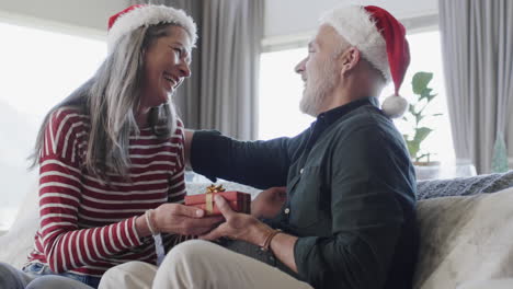 Middle-aged-caucasian-couple-in-santa-hats-gifting-at-christmas-at-home,-slow-motion