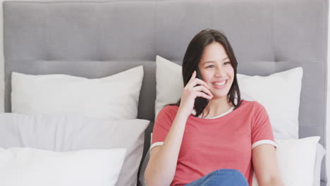 Happy-biracial-woman-sitting-on-bed-talking-on-smartphone,-copy-space,-slow-motion