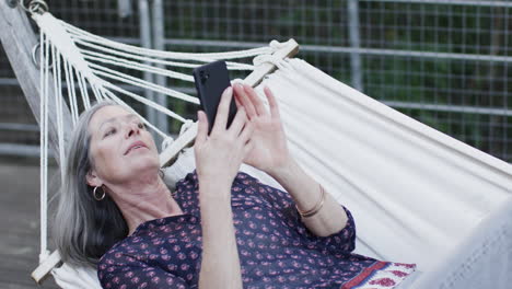 Caucasian-middle-aged-woman-using-smartphone-relaxing-in-hammock-on-terrace-in-nature,-slow-motion