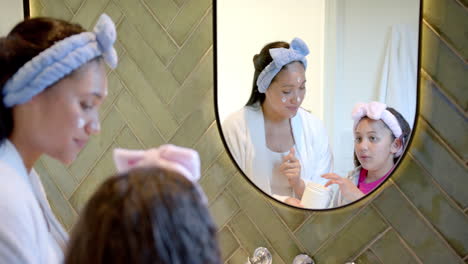 Happy-biracial-mother-and-daughter-with-headbands-putting-cream-on-faces-in-sunny-bathroom