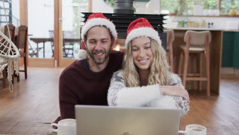 Vielfältiges-Paar-Mit-Weihnachtsmützen,-Das-Einen-Laptop-Für-Einen-Weihnachtsvideoanruf-In-Zeitlupe-Verwendet