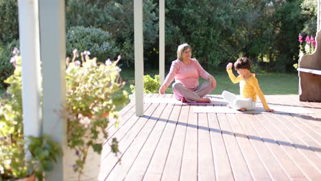 Happy-senior-biracial-grandmother-and-grandson-doing-yoga,-stretching,-slow-motion,-copy-space