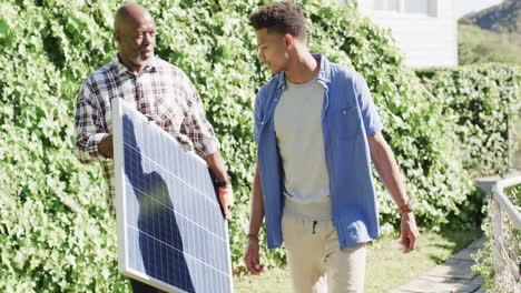 Padre-Afroamericano-Sosteniendo-Panel-Solar-Hablando-Con-Un-Hijo-Adulto-En-Un-Jardín-Soleado,-Cámara-Lenta