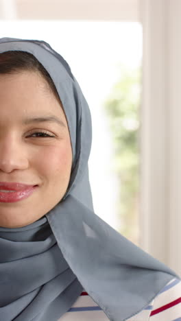 Vertical-video-of-happy-biracial-woman-in-hijab-smiling-to-camera-at-home,-copy-space,-slow-motion