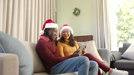 Happy-diverse-couple-in-christmas-hats-using-tablet-on-couch-at-home,-copy-space,-slow-motion