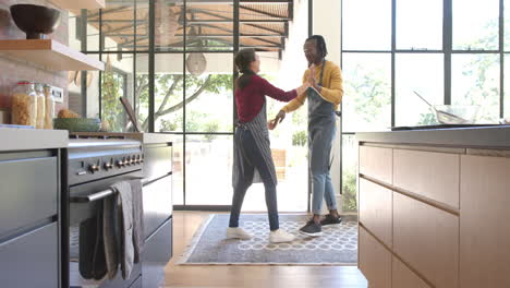 Happy-diverse-couple-in-aprons-dancing-in-sunny-kitchen,-copy-space,-slow-motion