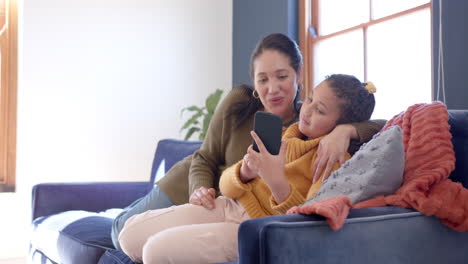 Happy-biracial-mother-and-daughter-embracing-on-sofa-and-using-smartphone-in-sunny-living-room