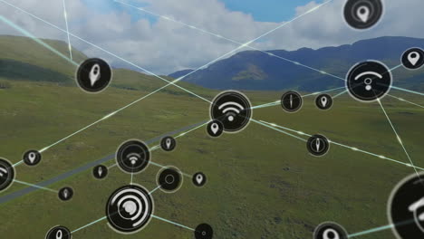 Animation-of-connected-icons-over-aerial-view-of-green-land-and-mountains-against-cloudy-sky