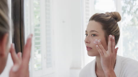 Biracial-woman-applying-face-cream-looking-at-mirror-in-bathroom,-slow-motion