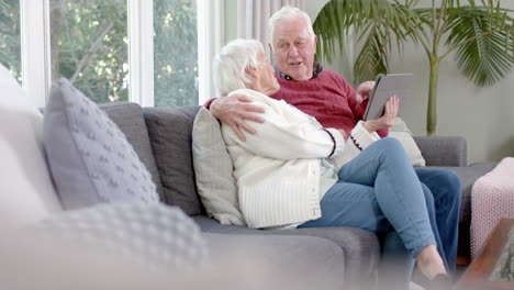 Happy-senior-caucasian-couple-sitting-on-sofa-using-tablet-at-home,-slow-motion,-copy-space