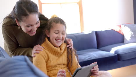 Happy-biracial-mother-and-daughter-sitting-on-sofa,-using-tablet-and-smiling-in-sunny-living-room