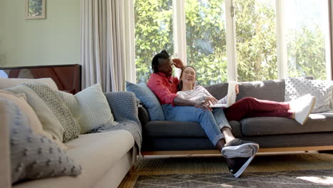 Happy-diverse-couple-relaxing-on-couch-using-laptop-in-living-room,-copy-space,-slow-motion