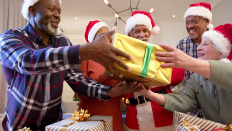 Happy-diverse-senior-friends-in-santa-outfit-and-christmas-hats-exchanging-gift-at-home,-slow-motion