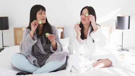 Happy-biracial-mother-and-adult-daughter-in-robes-putting-cucumber-slices-on-eyes,-slow-motion