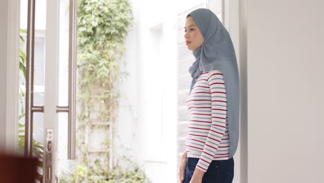 Portrait-of-biracial-woman-in-hijab-at-home-by-window-with-copy-space,-slow-motion