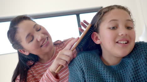 Happy-biracial-mother-brushing-daughter's-hair-in-sunny-bedroom
