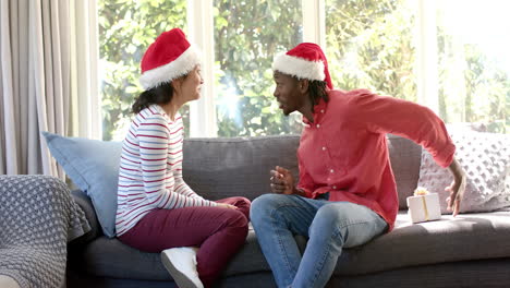 Glückliches,-Vielfältiges-Paar-Tauscht-Weihnachtsgeschenke-Auf-Der-Couch-Im-Sonnigen-Wohnzimmer-Aus,-Zeitlupe