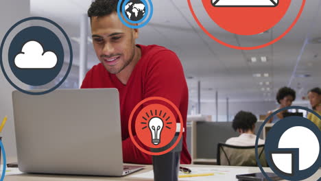 Animation-of-multiple-icons-over-smiling-biracial-man-standing-and-using-laptop-in-office