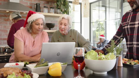 Happy-diverse-senior-female-friends-preparing-christmas-meal-using-laptop-in-kitchen,-slow-motion