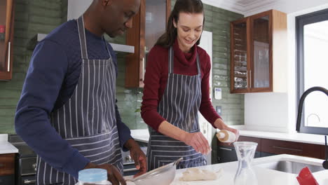 Happy-diverse-couple-standing-in-kitchen,-using-tablet-and-rolling-dough,slow-motion