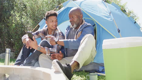 Glücklicher-Afroamerikanischer-Vater-Und-Erwachsener-Sohn-Sitzen-Draußen-Und-Benutzen-Ihr-Smartphone-In-Der-Sonne,-Zeitlupe