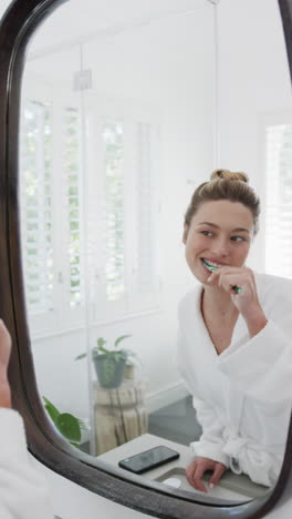 Vertical-video-of-biracial-woman-brushing-teeth-looking-in-mirror-in-bathroom,-slow-motion