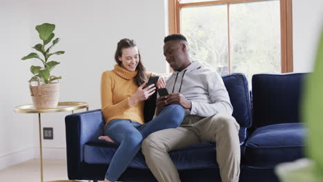 Happy-diverse-couple-sitting-on-sofa,-using-smartphone-and-credit-card-in-home,copy-space