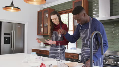 Happy-diverse-couple-standing-in-kitchen,-using-tablet-and-sprinkling-flour-on-worktop,slow-motion