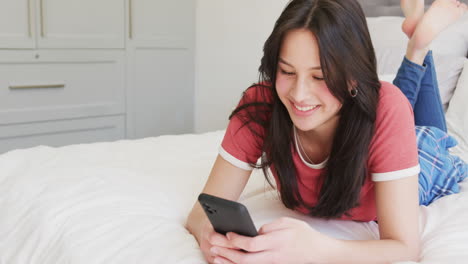 Happy-biracial-woman-lying-on-bed-using-smartphone,-copy-space,-slow-motion