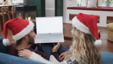 Vielfältiges-Paar-Mit-Weihnachtsmannmützen,-Das-Einen-Laptop-Mit-Kopierplatz-Auf-Dem-Bildschirm-In-Zeitlupe-Verwendet