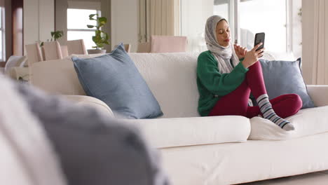 Biracial-woman-in-hijab-using-smartphone-on-sofa-at-home-with-copy-space,-slow-motion