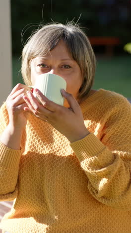 Vídeo-Vertical-De-Retrato-De-Una-Feliz-Mujer-Birracial-Mayor-Sosteniendo-Una-Taza-Fuera-De-Casa,-Cámara-Lenta