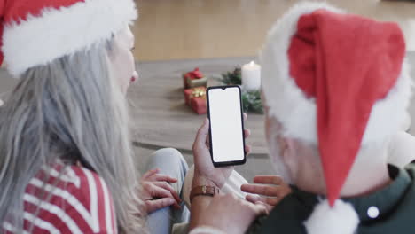Kaukasisches-Paar-In-Weihnachtsmützen-Smartphone-Mit-Leerem-Bildschirm-Zu-Weihnachten-Zu-Hause,-Zeitlupe