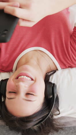 Vertical-video-of-happy-biracial-woman-in-headphones-lying-on-bed-using-smartphone,-slow-motion