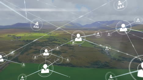Animation-of-connected-icons-over-aerial-view-of-green-land-against-mountains-and-cloudy-sky