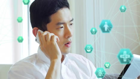 Animation-of-connected-icons-globes,-asian-man-looking-at-digital-tablet-and-talking-on-smartphone