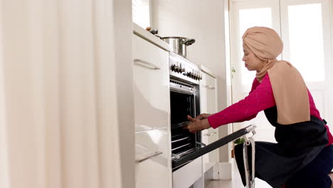 Biracial-woman-in-hijab-putting-tray-into-oven-in-kitchen-at-home-with-copy-space,-slow-motion