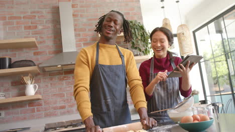 Happy-diverse-couple-in-aprons-using-tablet-and-baking-in-kitchen,-copy-space,-slow-motion