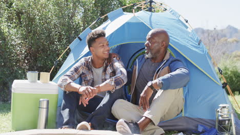 Happy-african-american-father-and-son-sitting-outside-tent-talking-in-the-sun,-slow-motion
