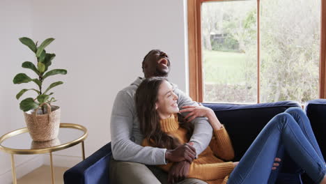 Happy-diverse-couple-sitting-on-sofa-laughing-and-embracing-in-home,copy-space