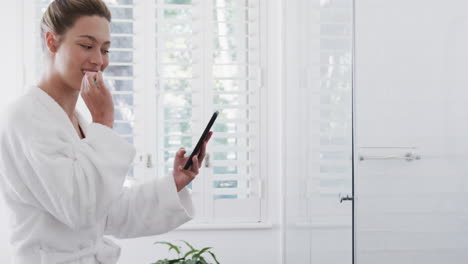 Happy-biracial-woman-brushing-teeth-using-smartphone-in-bathroom,-slow-motion