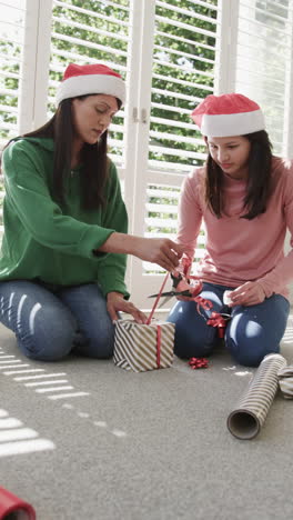 Vertical-video-of-happy-biracial-mother-and-daughter-in-christmas-hats-wrapping-gift,-slow-motion