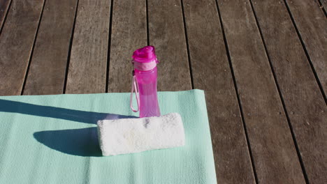 Water-bottle,-towel-and-yoga-mat-on-sunny-wooden-deck-outdoors,-copy-space,-slow-motion