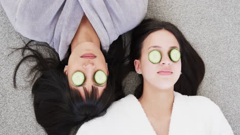 Happy-biracial-mother-and-adult-daughter-in-robes-lying-with-cucumber-slices-on-eyes,-slow-motion