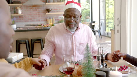 Happy-african-american-grandfather-holding-hands-with-family-saying-grace-at-christmas-dinner-table