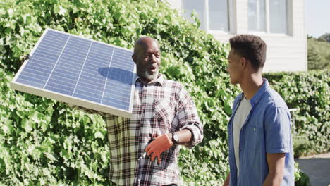 Happy-african-american-father-with-solar-panel-talking-to-adult-son-in-sunny-garden,-slow-motion