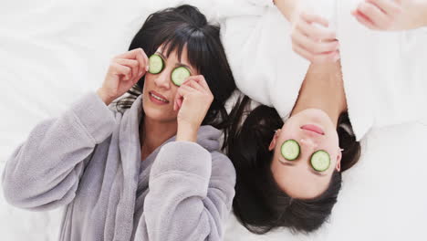 Happy-biracial-mother-and-adult-daughter-taking-cucumber-slices-from-eyes-and-laughing,-slow-motion